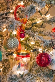 A festive Christmas tree adorned with various colorful ornaments, including a red ball, a green and gold textured ornament, and a small glass jar with a decorative scene inside. The branches are dusted with artificial snow and illuminated by warm, white string lights, creating a cozy and joyful atmosphere.