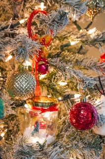 A festive Christmas tree adorned with various colorful ornaments, including a red ball, a green and gold textured ornament, and a small glass jar with a decorative scene inside. The branches are dusted with artificial snow and illuminated by warm, white string lights, creating a cozy and joyful atmosphere.