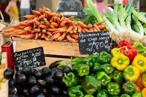 A vibrant display of fresh vegetables at a market stall, including stacks of carrots, eggplants, bell peppers, and leeks. Handwritten chalkboard signs indicate the names and prices of the produce.