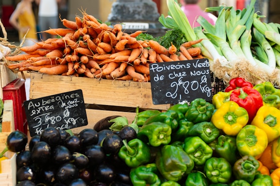 A vibrant display of fresh vegetables at a market stall, including stacks of carrots, eggplants, bell peppers, and leeks. Handwritten chalkboard signs indicate the names and prices of the produce.