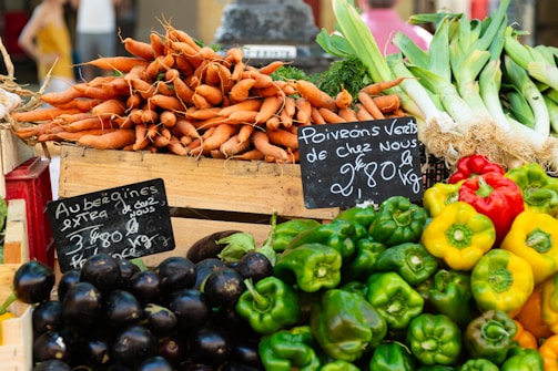 A vibrant display of fresh vegetables at a market stall, including stacks of carrots, eggplants, bell peppers, and leeks. Handwritten chalkboard signs indicate the names and prices of the produce.