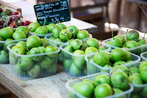 Several containers filled with fresh green pears are arranged on a wooden table at a market. A sign displays information about the pears in French, with text written on a small chalkboard. To the side, there are a few strawberries in a separate container adding a touch of red color.