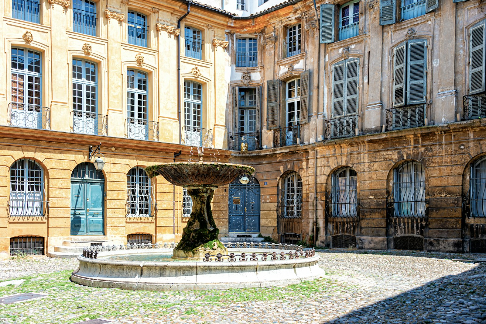 A tiered stone fountain in a cobbled square in Aix-en-Provence, framed by golden façades with shutters and wrought-iron balconies