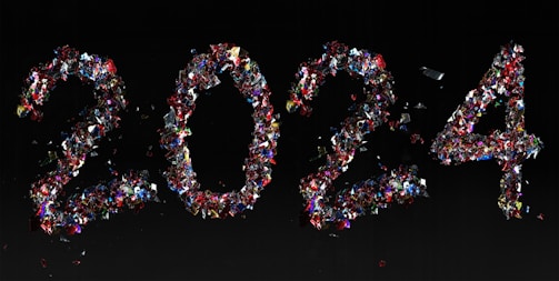 The numbers '2024' are creatively formed using an assortment of colorful, reflective confetti on a dark background. The confetti pieces are predominantly red, blue, and silver, creating a vibrant and festive composition.