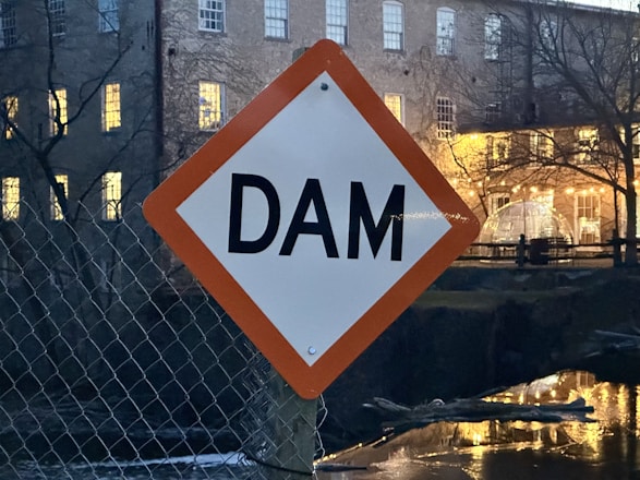 A diamond-shaped sign with the word 'DAM' is mounted on a fence in the foreground. In the background, there is a building with several windows, some of which are lit up, and a tree with leafless branches. The scene is set near a body of water, reflecting light from the building.