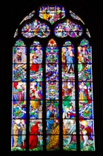 An intricately designed stained glass window featuring multiple detailed panels depicting scenes with various figures, likely of religious significance. The artwork includes rich patterns, vibrant colors, and displays a range of activities and interactions among the figures, surrounded by ornate floral and geometric designs.
