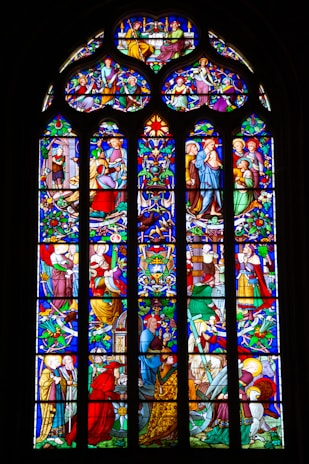 An intricately designed stained glass window featuring multiple detailed panels depicting scenes with various figures, likely of religious significance. The artwork includes rich patterns, vibrant colors, and displays a range of activities and interactions among the figures, surrounded by ornate floral and geometric designs.