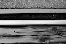 A close-up view of a railroad track consisting of a metal rail, wooden sleeper, and gravel ballast. The textures of the gravel, wood grain, and metal are distinctly visible.