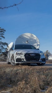 A sleek gray Audi car is parked on a road with large satellite dishes in the background. The setting seems to be outdoors with clear blue sky and dry grass in the foreground.