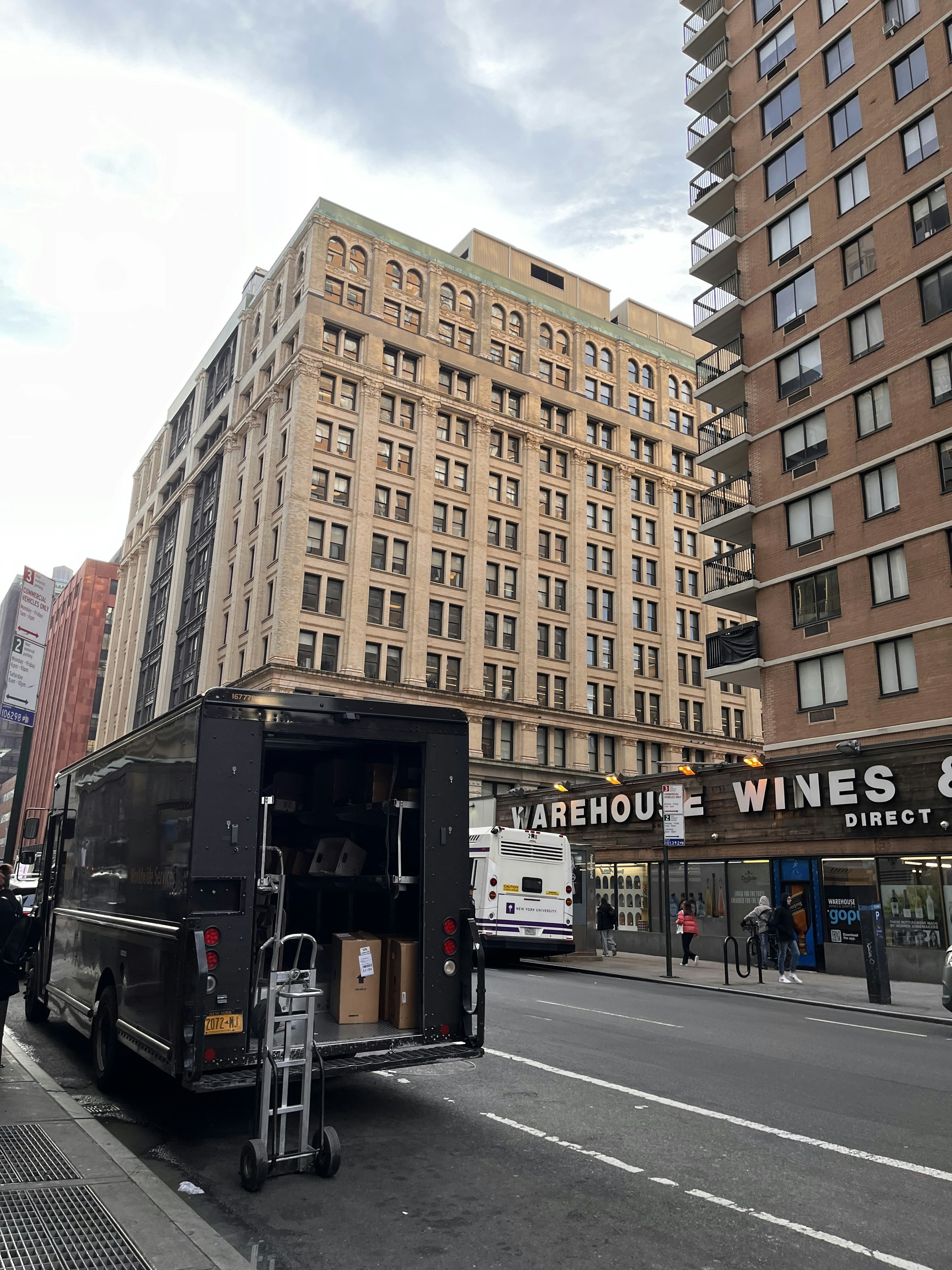 A large delivery truck is parked on the side of a city street with its back door open, revealing boxes and a dolly. In the background, tall buildings with numerous windows rise, and a sign for Warehouse Wines is visible. A city bus is stopped nearby, with several pedestrians walking on the sidewalk.