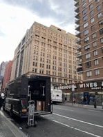 A large delivery truck is parked on the side of a city street with its back door open, revealing boxes and a dolly. In the background, tall buildings with numerous windows rise, and a sign for Warehouse Wines is visible. A city bus is stopped nearby, with several pedestrians walking on the sidewalk.