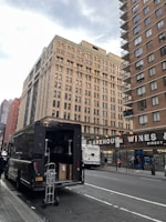 A large delivery truck is parked on the side of a city street with its back door open, revealing boxes and a dolly. In the background, tall buildings with numerous windows rise, and a sign for Warehouse Wines is visible. A city bus is stopped nearby, with several pedestrians walking on the sidewalk.