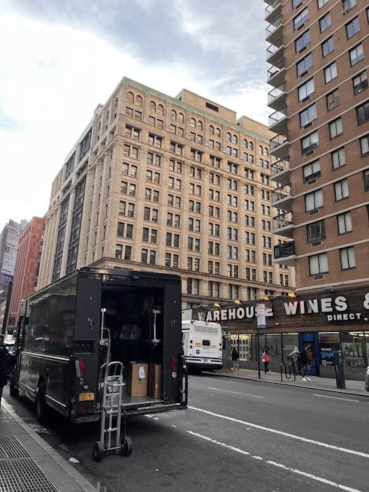 A large delivery truck is parked on the side of a city street with its back door open, revealing boxes and a dolly. In the background, tall buildings with numerous windows rise, and a sign for Warehouse Wines is visible. A city bus is stopped nearby, with several pedestrians walking on the sidewalk.