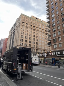 A large delivery truck is parked on the side of a city street with its back door open, revealing boxes and a dolly. In the background, tall buildings with numerous windows rise, and a sign for Warehouse Wines is visible. A city bus is stopped nearby, with several pedestrians walking on the sidewalk.