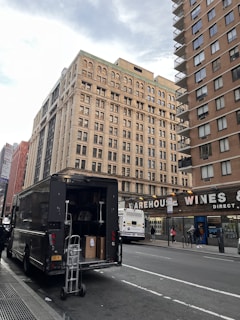 A large delivery truck is parked on the side of a city street with its back door open, revealing boxes and a dolly. In the background, tall buildings with numerous windows rise, and a sign for Warehouse Wines is visible. A city bus is stopped nearby, with several pedestrians walking on the sidewalk.