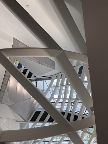 Various intersecting and overlapping structural beams and geometric shapes, featuring steel or concrete materials. The architecture creates a complex interplay of lines and shadows, with some natural light filtering in from the windows in the background.