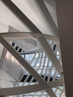 Various intersecting and overlapping structural beams and geometric shapes, featuring steel or concrete materials. The architecture creates a complex interplay of lines and shadows, with some natural light filtering in from the windows in the background.