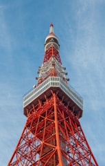 A tall red and white lattice communications tower set against a blue sky. The structure features intricate geometric patterns and metal framework with a large observation deck located near the top.