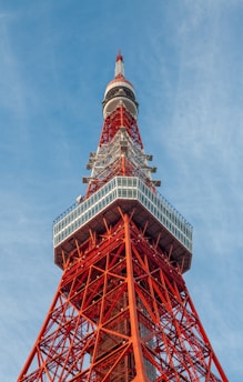 A tall red and white lattice communications tower set against a blue sky. The structure features intricate geometric patterns and metal framework with a large observation deck located near the top.