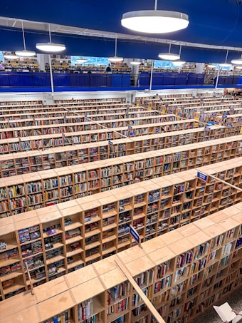 A large, multi-level library with numerous rows of wooden bookshelves filled with a wide variety of books. The image captures a top view, showcasing the extensive organization and arrangement of the shelves. Bright round ceiling lights illuminate the space, and a balcony level with a blue railing is visible above. Some people are seen browsing through the books on the upper level.