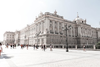 A grand, historical palace with intricate architectural details and numerous large windows. The building has statues along its ledge and is accompanied by traditional street lamps. People are walking in the spacious courtyard, suggesting it is a popular tourist destination.