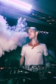 A person wearing a cap and a white shirt stands behind DJ equipment, with a microphone in the foreground. They are exhaling a large cloud of vapor or smoke. The setting appears to be a dark indoor venue with colorful lighting, giving a nightlife atmosphere.