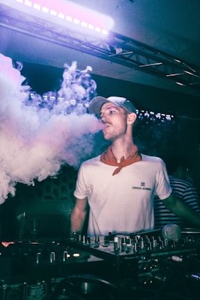 A person wearing a cap and a white shirt stands behind DJ equipment, with a microphone in the foreground. They are exhaling a large cloud of vapor or smoke. The setting appears to be a dark indoor venue with colorful lighting, giving a nightlife atmosphere.
