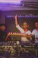 A group of people are in a lively, dimly lit room, likely a club or party setting. One person is holding a tray of pink drinks and raising his hand in excitement. They are dressed festively, with one wearing a straw hat and sunglasses. A DJ setup with Pioneer DJ equipment is visible in the foreground.
