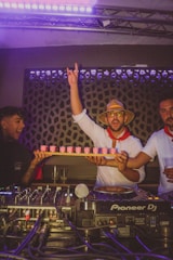 A group of people are in a lively, dimly lit room, likely a club or party setting. One person is holding a tray of pink drinks and raising his hand in excitement. They are dressed festively, with one wearing a straw hat and sunglasses. A DJ setup with Pioneer DJ equipment is visible in the foreground.