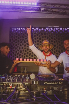 A group of people are in a lively, dimly lit room, likely a club or party setting. One person is holding a tray of pink drinks and raising his hand in excitement. They are dressed festively, with one wearing a straw hat and sunglasses. A DJ setup with Pioneer DJ equipment is visible in the foreground.
