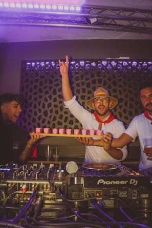 A group of people are in a lively, dimly lit room, likely a club or party setting. One person is holding a tray of pink drinks and raising his hand in excitement. They are dressed festively, with one wearing a straw hat and sunglasses. A DJ setup with Pioneer DJ equipment is visible in the foreground.