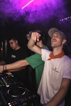 Three individuals are in a dark setting, likely in a club or music venue. One person at the forefront is wearing a light-colored cap and a white shirt with a red bandana around the neck, seemingly engaging with the crowd or the camera. Another person wearing a green shirt is partially obscured, and a third individual in a black shirt with 'Tupac' written on it is focusing on a DJ mixer with colorful lights. There is purple ambient lighting and smoke in the background, suggesting a lively atmosphere.