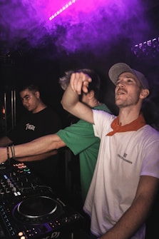 Three individuals are in a dark setting, likely in a club or music venue. One person at the forefront is wearing a light-colored cap and a white shirt with a red bandana around the neck, seemingly engaging with the crowd or the camera. Another person wearing a green shirt is partially obscured, and a third individual in a black shirt with 'Tupac' written on it is focusing on a DJ mixer with colorful lights. There is purple ambient lighting and smoke in the background, suggesting a lively atmosphere.