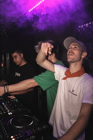 Three individuals are in a dark setting, likely in a club or music venue. One person at the forefront is wearing a light-colored cap and a white shirt with a red bandana around the neck, seemingly engaging with the crowd or the camera. Another person wearing a green shirt is partially obscured, and a third individual in a black shirt with 'Tupac' written on it is focusing on a DJ mixer with colorful lights. There is purple ambient lighting and smoke in the background, suggesting a lively atmosphere.