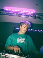 A person wearing a teal shirt with the word 'Supreme' on it stands behind a DJ mixer. They have headphones on and are under a set of purple stage lights, suggesting they are performing or mixing music.