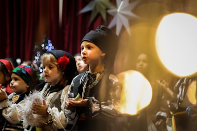 A group of children dressed in traditional clothing, possibly for a cultural or festive performance. They are standing on stage, illuminated by warm lighting, and appear to be singing or reciting something. The setting includes decorative elements, such as large star shapes and strings of lights in the background.