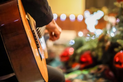 A close-up view of a person's hand playing an acoustic guitar, with festive holiday decorations blurred in the background. Soft, warm lighting enhances the cozy atmosphere.