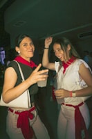 Two women dressed in casual white and red clothing are enjoying a night out, holding drinks and smiling. The atmosphere suggests a festive or party environment, with dim lighting and other indistinct figures in the background.