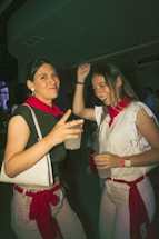 Two women dressed in casual white and red clothing are enjoying a night out, holding drinks and smiling. The atmosphere suggests a festive or party environment, with dim lighting and other indistinct figures in the background.