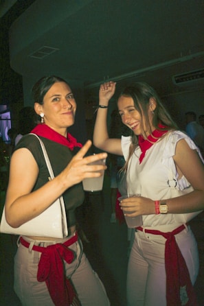 Two women dressed in casual white and red clothing are enjoying a night out, holding drinks and smiling. The atmosphere suggests a festive or party environment, with dim lighting and other indistinct figures in the background.