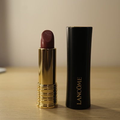 A dark red lipstick with a gold casing is positioned upright on a wooden surface. Next to it is the black and gold cap with the brand name Lancôme visible. The background is softly blurred in neutral tones.