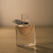 A glass perfume bottle with a transparent square cap and a dark ribbon near the neck. It is filled with a light pinkish liquid. The bottle is placed on a wooden surface with a warm, soft light creating a serene ambiance.