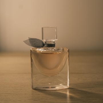 A glass perfume bottle with a transparent square cap and a dark ribbon near the neck. It is filled with a light pinkish liquid. The bottle is placed on a wooden surface with a warm, soft light creating a serene ambiance.