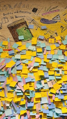 A colorful wall filled with numerous sticky notes of various colors, predominantly yellow, blue, and pink. Above the notes, there is a mural featuring drawings of books, musical notes, and other stationery items. The text 'Books, and Everything in Between' is visible within the mural.