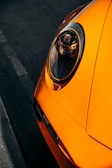 Close-up of an orange car's headlight in a dimly lit setting. The headlight design is modern and sleek, with reflective surfaces that catch the light. The car's bold orange color contrasts with the dark asphalt.