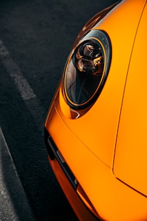 Close-up of an orange car's headlight in a dimly lit setting. The headlight design is modern and sleek, with reflective surfaces that catch the light. The car's bold orange color contrasts with the dark asphalt.