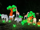 A vibrant night scene featuring illuminated animal sculptures in a park setting. Giraffes, zebras, and lions are surrounded by bright green artificial foliage and trees, creating a vivid display against the dark background.
