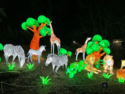 A vibrant night scene featuring illuminated animal sculptures in a park setting. Giraffes, zebras, and lions are surrounded by bright green artificial foliage and trees, creating a vivid display against the dark background.