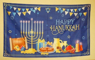 A festive Hanukkah tapestry featuring a lit menorah with nine candles, surrounded by traditional items like dreidels, donuts, a gift box, coins, and a shofar. A pair of lit candles, a pomegranate, and a Torah scroll add to the scene. 'Happy Hanukkah' is prominently displayed in decorative lettering. The background is deep blue with a border and scattered Star of David motifs, accompanied by bunting flags in blue, white, and yellow.