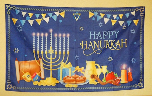 A festive Hanukkah tapestry featuring a lit menorah with nine candles, surrounded by traditional items like dreidels, donuts, a gift box, coins, and a shofar. A pair of lit candles, a pomegranate, and a Torah scroll add to the scene. 'Happy Hanukkah' is prominently displayed in decorative lettering. The background is deep blue with a border and scattered Star of David motifs, accompanied by bunting flags in blue, white, and yellow.